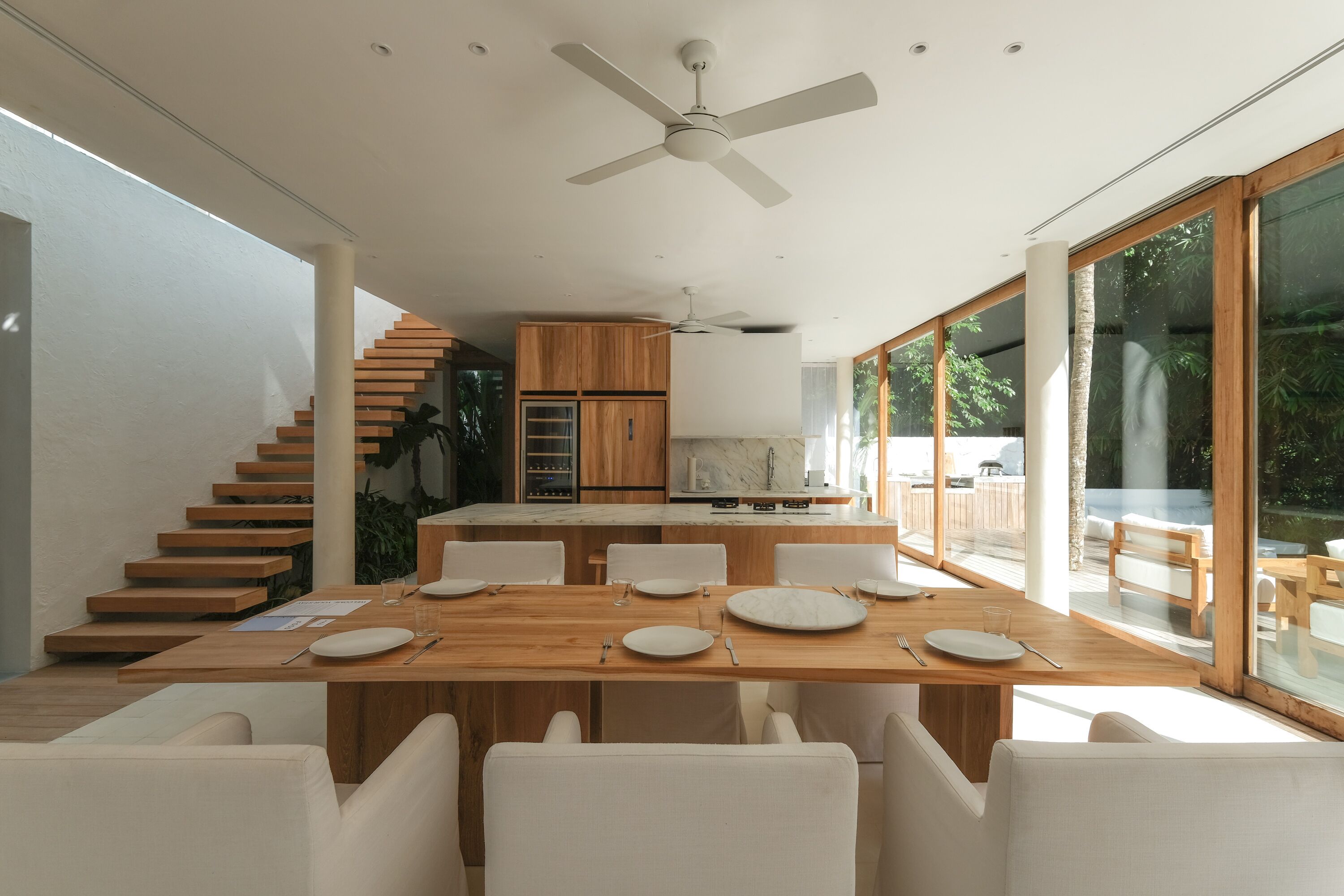 A modern dining area with a wooden table set for four, white chairs, and a kitchen in the background with wooden cabinets and a marble countertop.