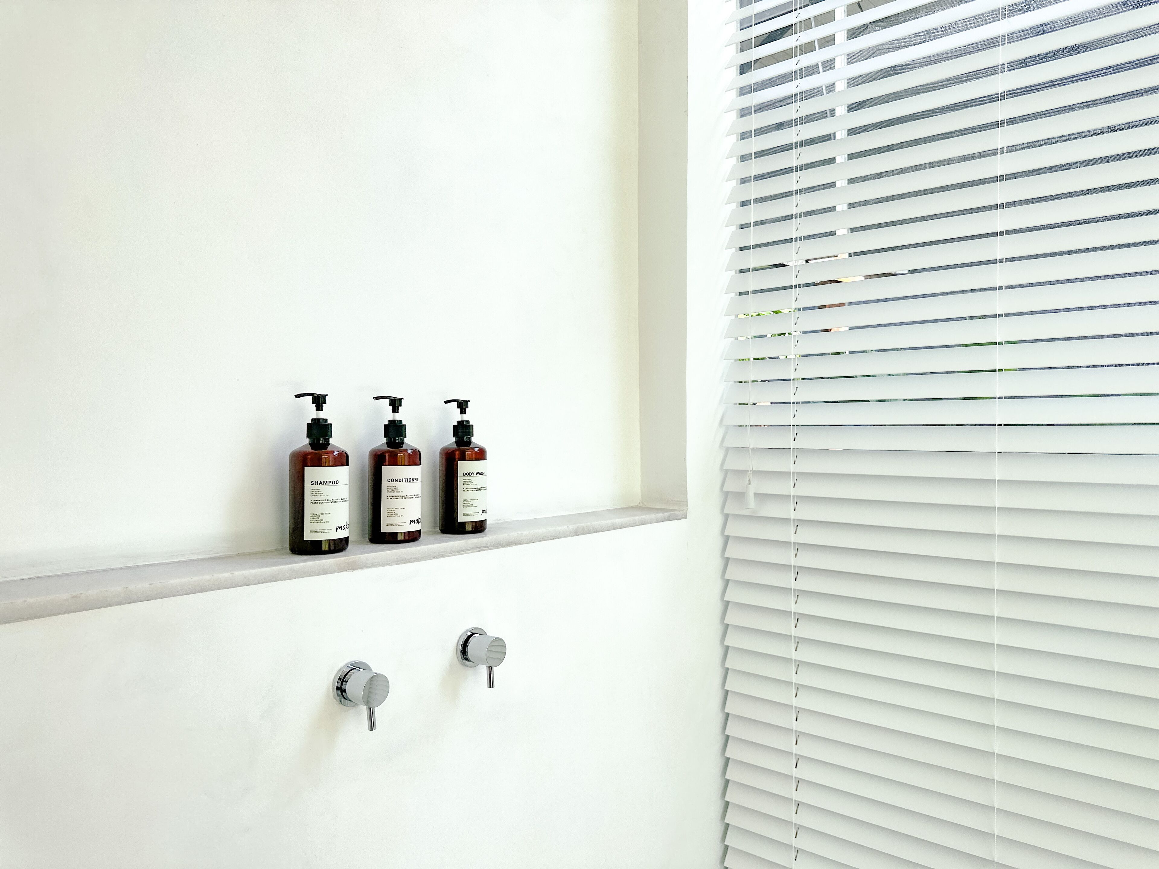 A bathroom with three bottles of shampoo, conditioner, and body wash on a shelf, and two faucets below.