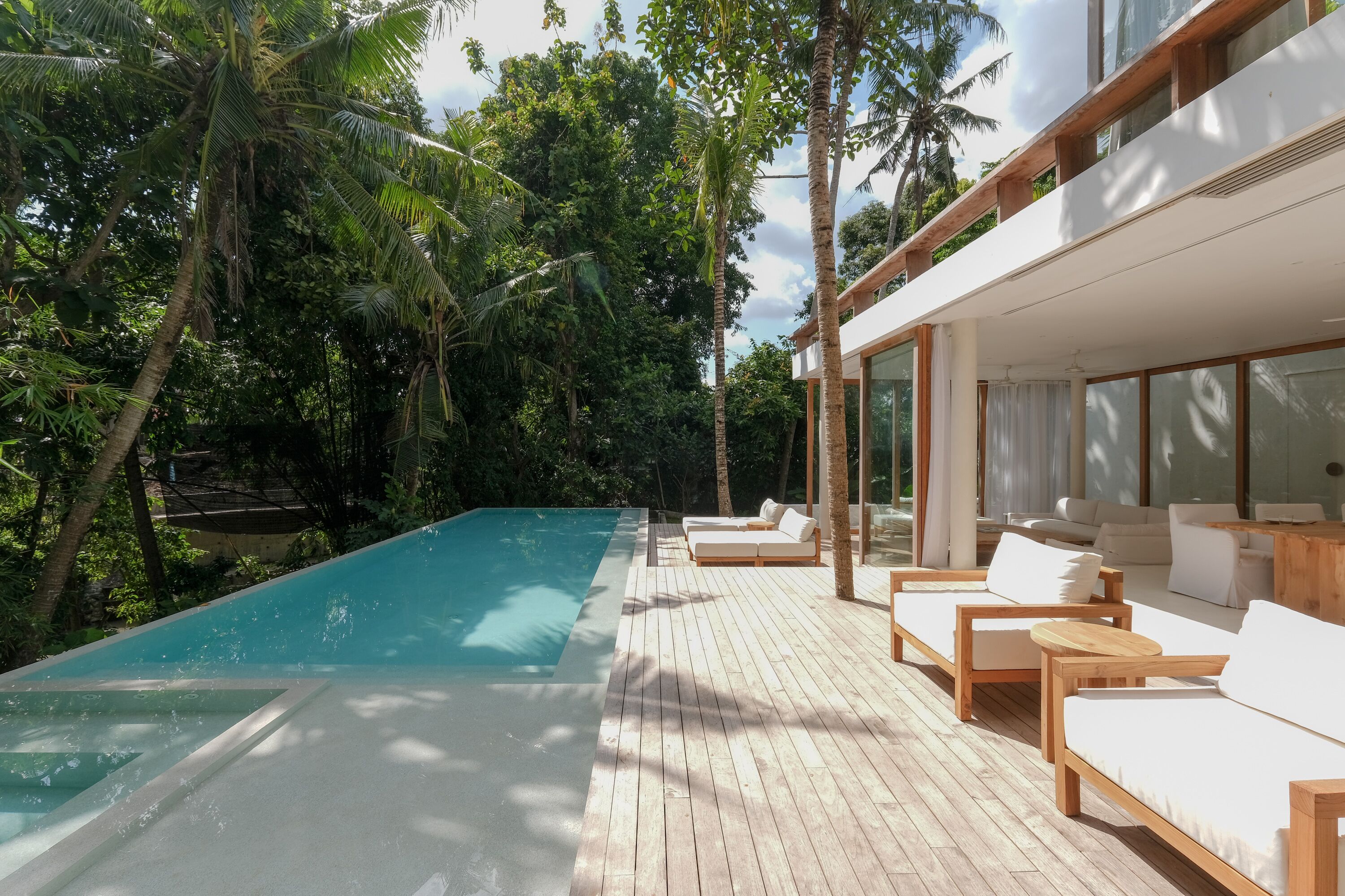 A modern outdoor pool area with wooden deck and lounge chairs, surrounded by lush greenery.