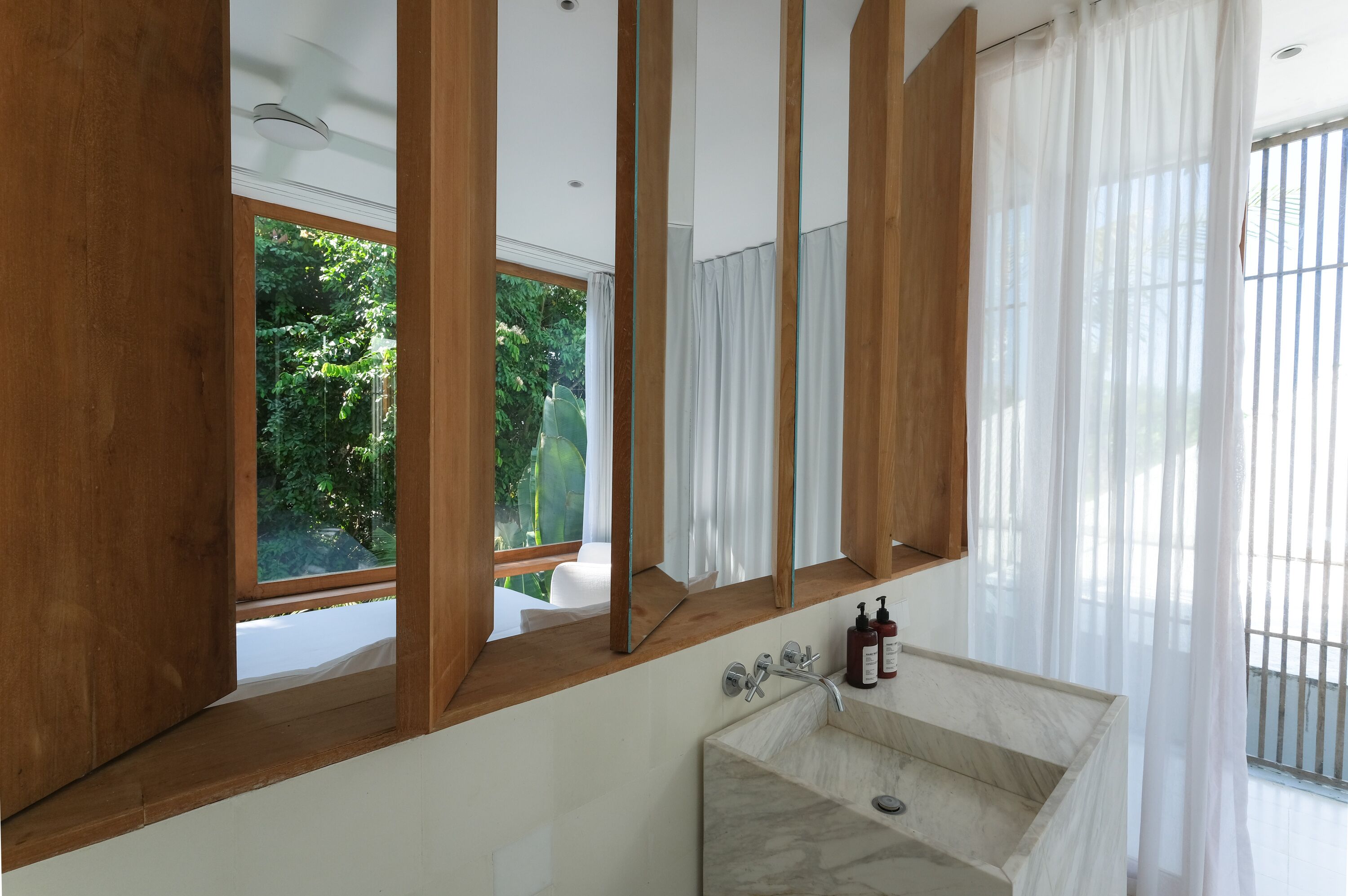 A modern bathroom with a large mirror, wooden beams, a marble sink, and a view of greenery outside.