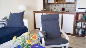 Ein Wohnzimmer mit Sofa, einem Sessel, einem kleinen Tisch mit Blumen sowie einer Küchenzeile mit Schränken und Geräten.