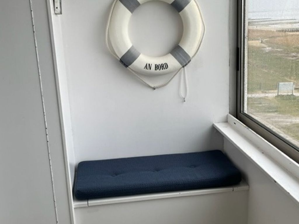 Ein Fensterplatz mit dunklem Kissen und einem Rettungsring an der Wand.