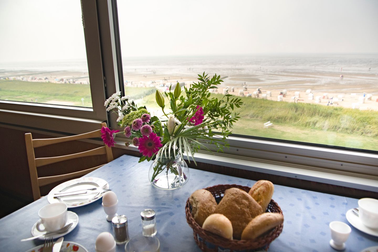 Una mesa de comedor con vista a la playa, un jarrón con flores y una cesta de pan.