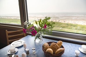 Een eettafel met uitzicht op het strand, een vaas met bloemen en een mand met brood.