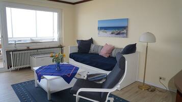Ein gemütliches Zimmer mit Bett, Tisch mit Blumenvase, Stuhl, Stehlampe und Fenster mit Meerblick.