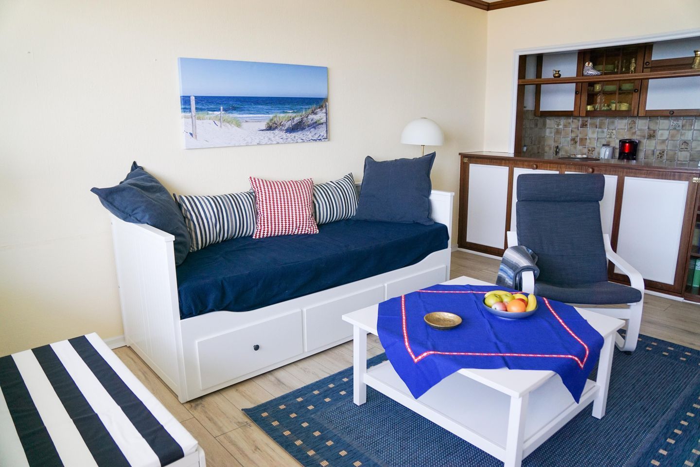 Un salon avec un canapé, une table à manger agrémentée d’un panier de fruits, un coin cuisine et un tableau représentant une vue sur la plage.