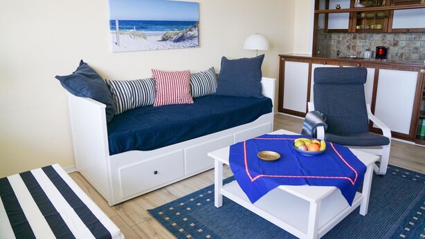 A living room with a sofa, a dining table with a fruit bowl, a kitchen area, and a beach view painting. - Ferienwohnung mit Maritimer Ausstattung, Seeblick und Schwimmbad - Haus Seehund (Cuxhaven)