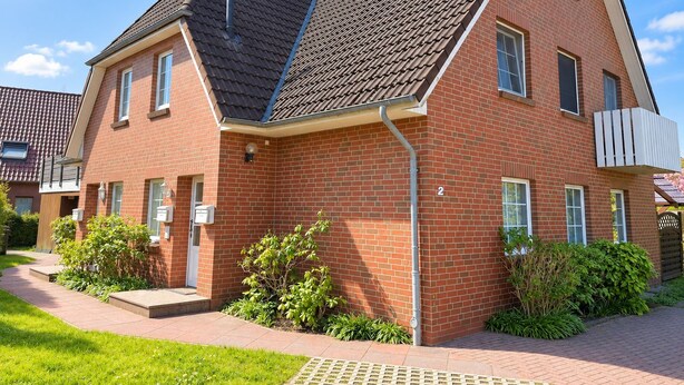A two-story brick house with a sloped roof, a visible number '2' on the side, and a small garden area with shrubs.