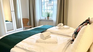 Ett hotellrum med två sängar, ett fönster med gardiner, en stol och ett litet bord med en växt.