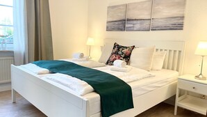A neatly made bed with a green throw, white bedding, and a floral pillow. There are two bedside tables with lamps, a window with curtains, and a wall with three paintings.