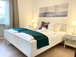 A neatly made bed with a green throw, white bedding, and a floral pillow. There are two bedside tables with lamps, a window with curtains, and a wall with three paintings.