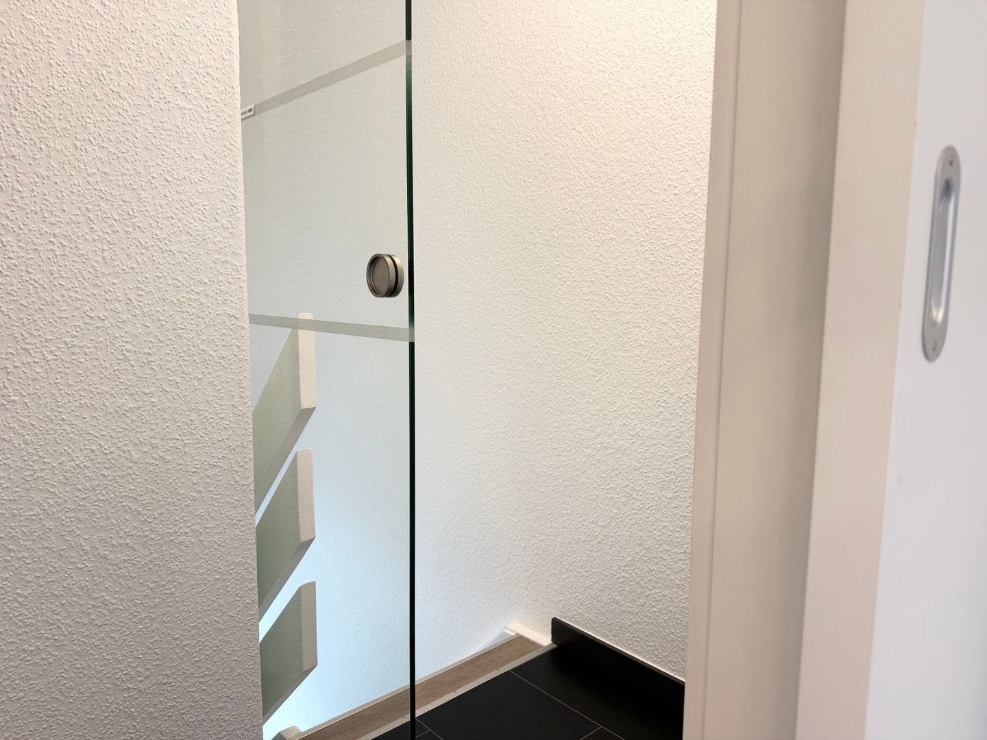 A modern interior with a glass door, white textured walls, and a visible staircase.