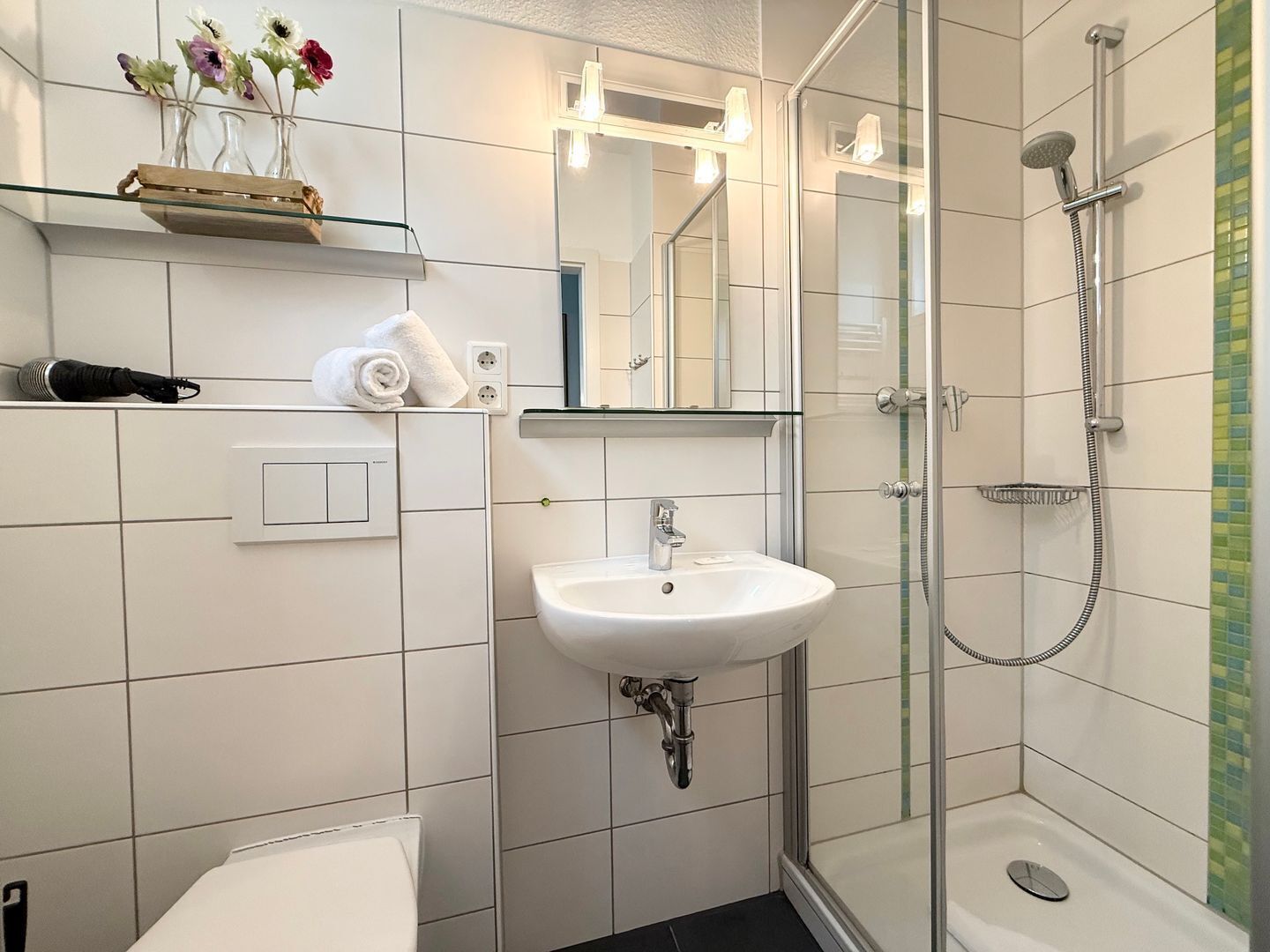 Ein Badezimmer mit weißem Waschbecken, einer Duschkabine aus Glas, einem Spiegel und einem Regal mit Blumen.