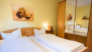 A double bed with white linens, a wooden headboard, a bedside table with a lamp, a mirror, and a painting on the wall.