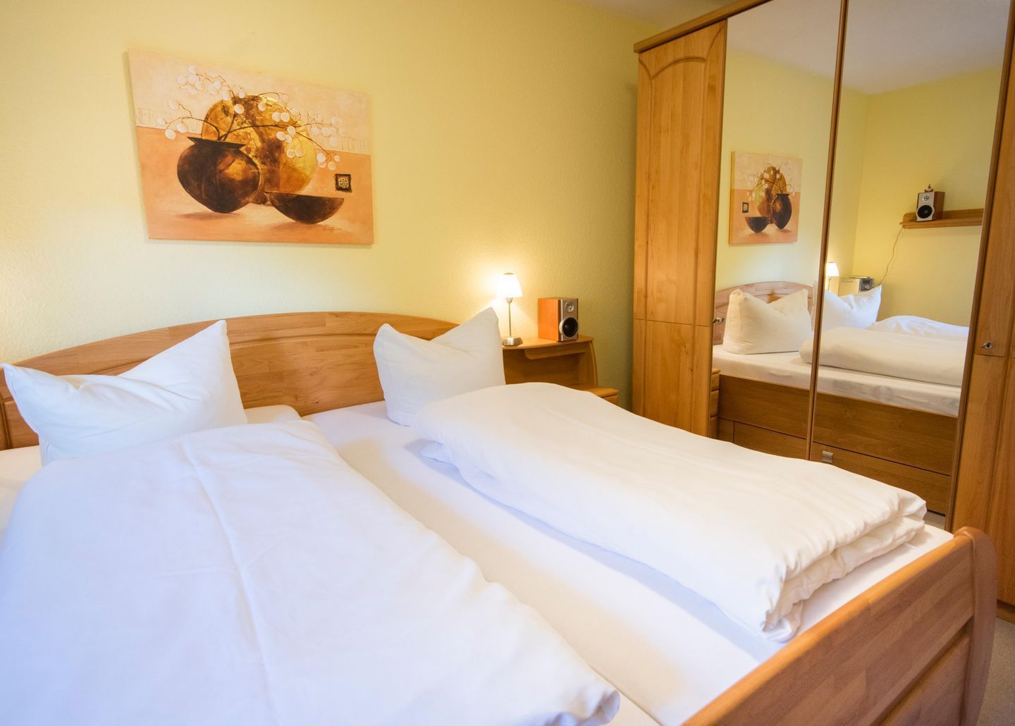 Ein Doppelbett mit weißen Leinentüchern, ein hölzerner Kopfseitenverkleidung, ein Nachttisch mit Lampe, ein Spiegel und ein Bild an der Wand.