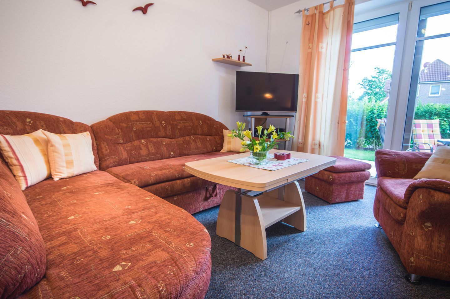 Ein Wohnzimmer mit einer Couch, einem kleinen Tisch mit Blumen, einem Fernseher und Blick ins Freie.