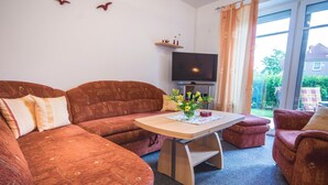A living room with a sofa, a small table with a vase of flowers, a television, and a view of the outside.