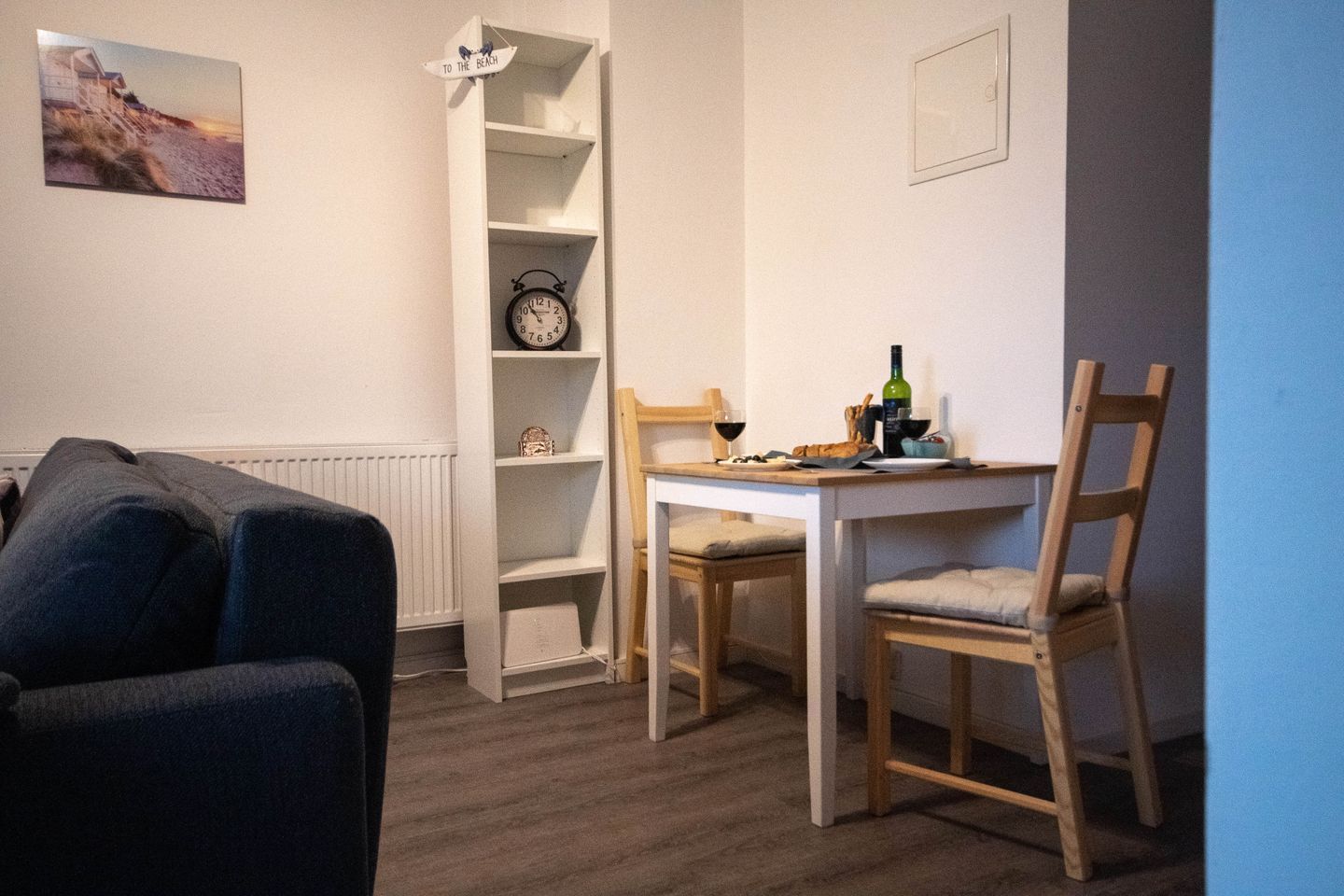 A room with a white wall, a clock, a small table with chairs, a radiator, and a picture of a beach.