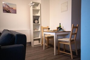 A room with a white wall, a clock, a small table with chairs, a radiator, and a picture of a beach.