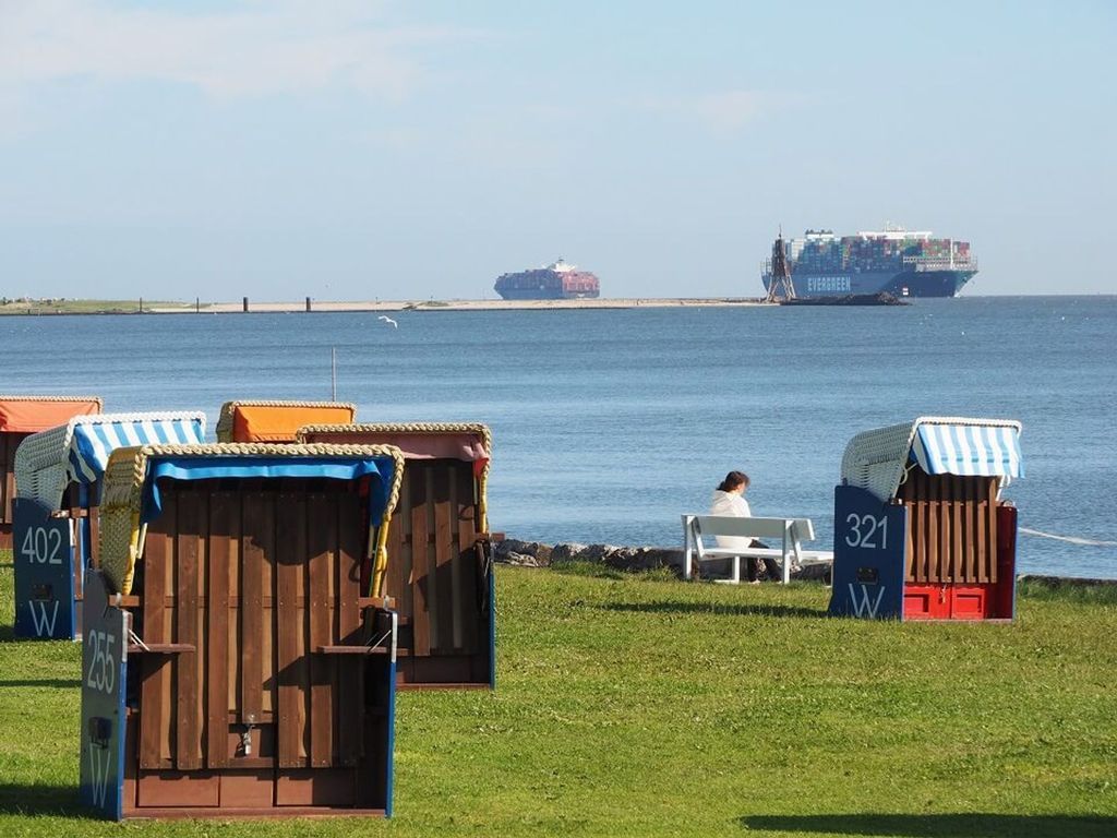 Beachside wooden structures with numbers, a person sitting on a bench, and a cargo ship in the background.