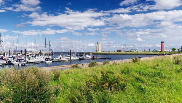 A marina with multiple sailboats docked, a lighthouse, and industrial structures in the background.