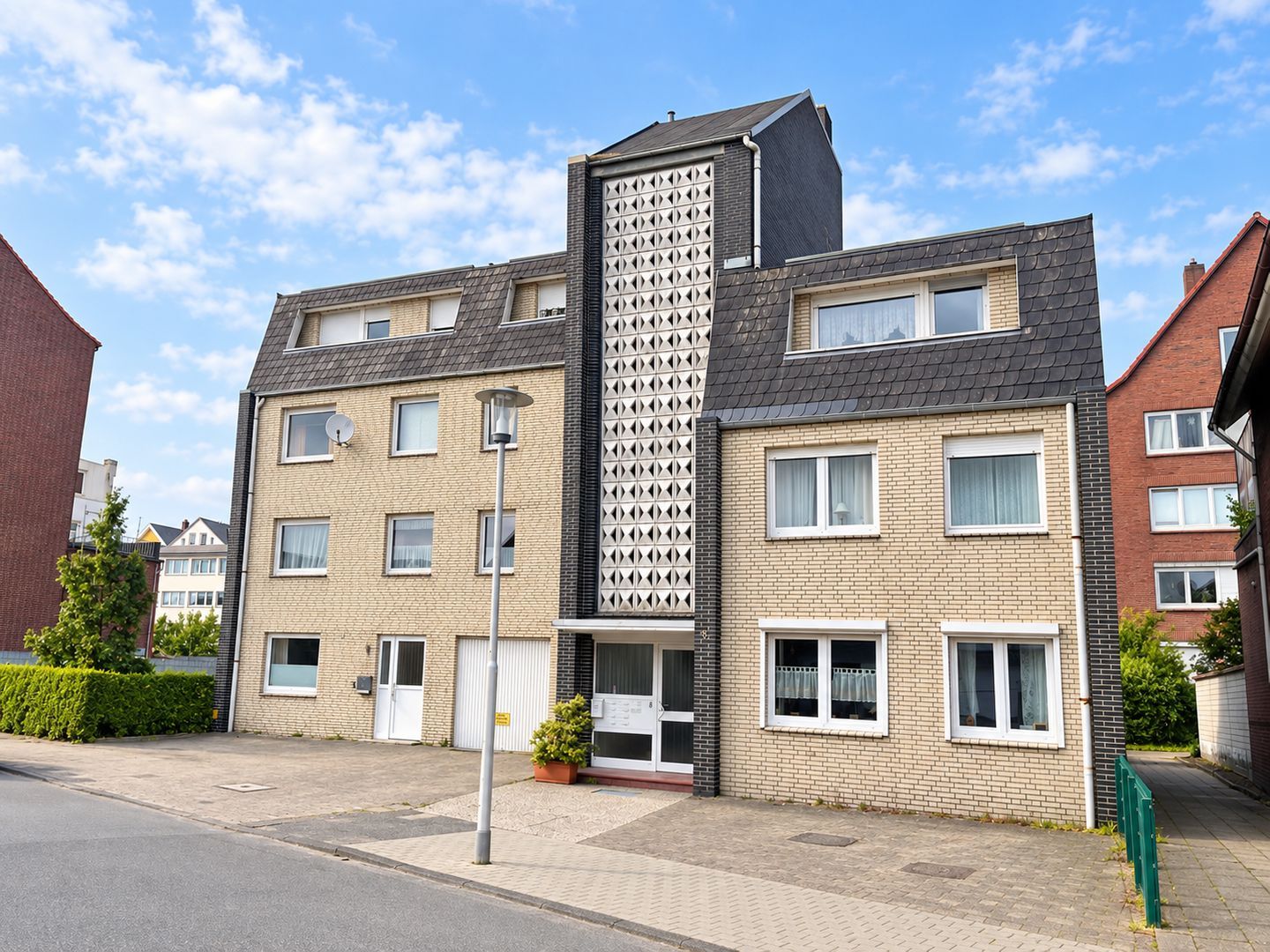 Ein mehrstöckiges Wohngebäude mit einem zentralen Turm, zahlreichen Fenstern und einem gepflasterten Eingangsbereich.