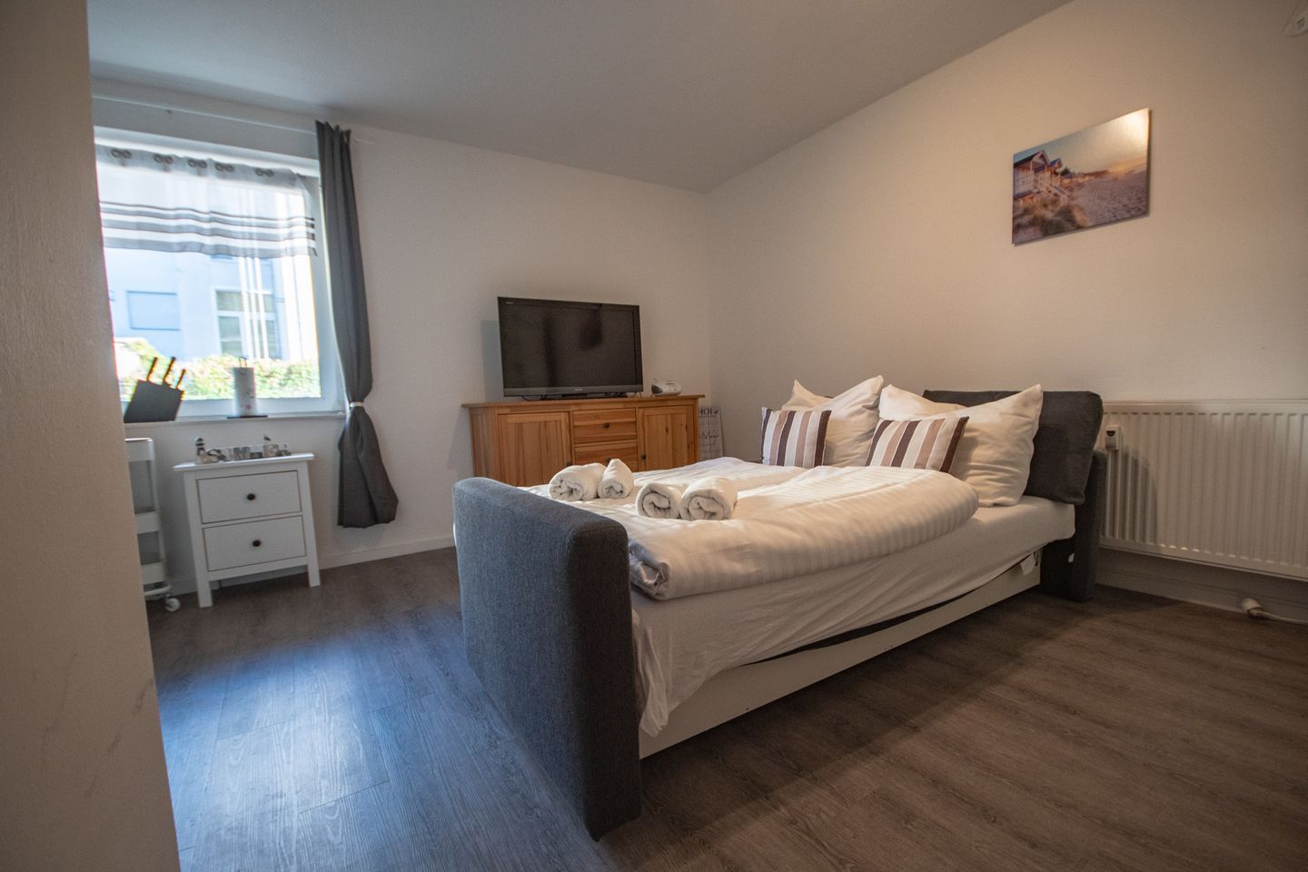 Ein Schlafzimmer mit Bett, Fernseher, Kommode und einem Fenster mit Vorhängen.