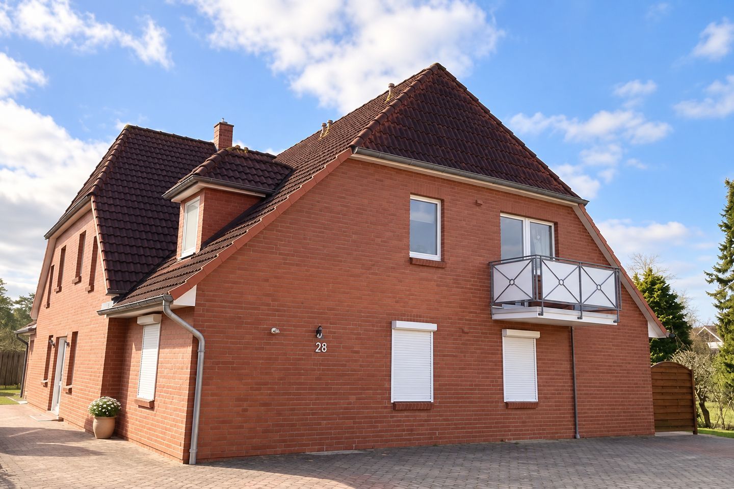 Ein zweigeschossiges Backsteinhäuschen mit Balkon und Hausnummer 28, sowie einer gepflasterten Auffahrt.