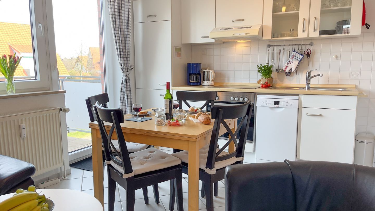 Una cocina moderna con armarios blancos, una cafetera y un lavavajillas. Hay una mesa de comedor con sillas y una ventana con vistas a edificios.