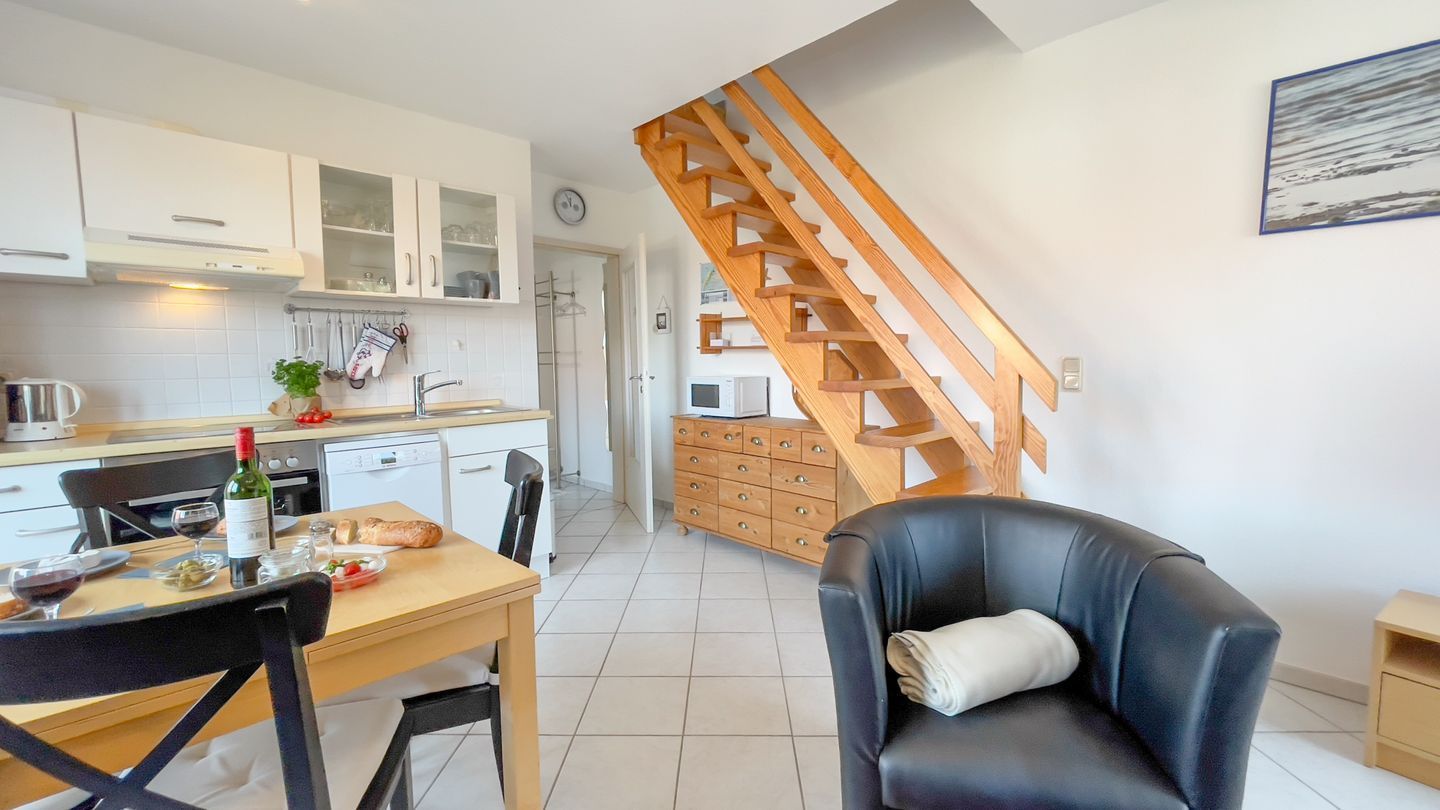 Een moderne keuken met witte kasten, een houten trap, een zithoek met een tafel en stoelen, en een zwarte leren fauteuil.