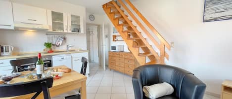 Une cuisine moderne avec des meubles blancs, un escalier en bois, un coin repas avec une table et des chaises, et un fauteuil en cuir noir.