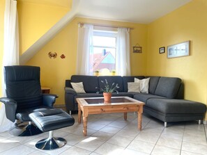 A living room with a black leather chair, a grey sofa, a wooden coffee table, and a window with white curtains.