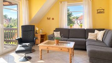 Ein Wohnzimmer mit einer Couch, einem Sessel und einem Couchtisch, ein Balkon mit Blick und einer gelben Wand.