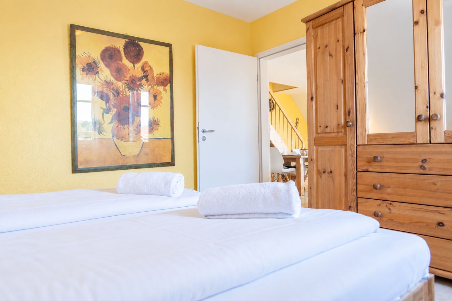 Quarto de hotel com duas camas, um guarda-roupa de madeira, um quadro de girassóis e uma porta branca que dá para uma escadaria.