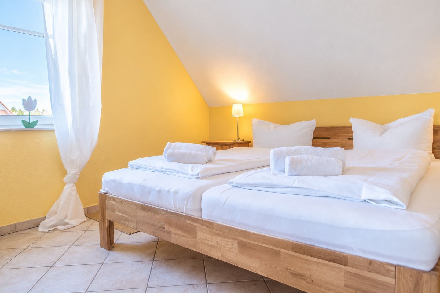 Ein Doppelbett mit weißen Laken, ein Nachttisch mit Lampe und ein Fenster mit Vorhängen.