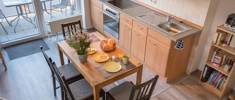 Cocina compacta con armarios de madera, microondas integrado, estufa y lavamanos. Cuenta con una mesa de comedor con sillas y una pequeña planta en maceta.