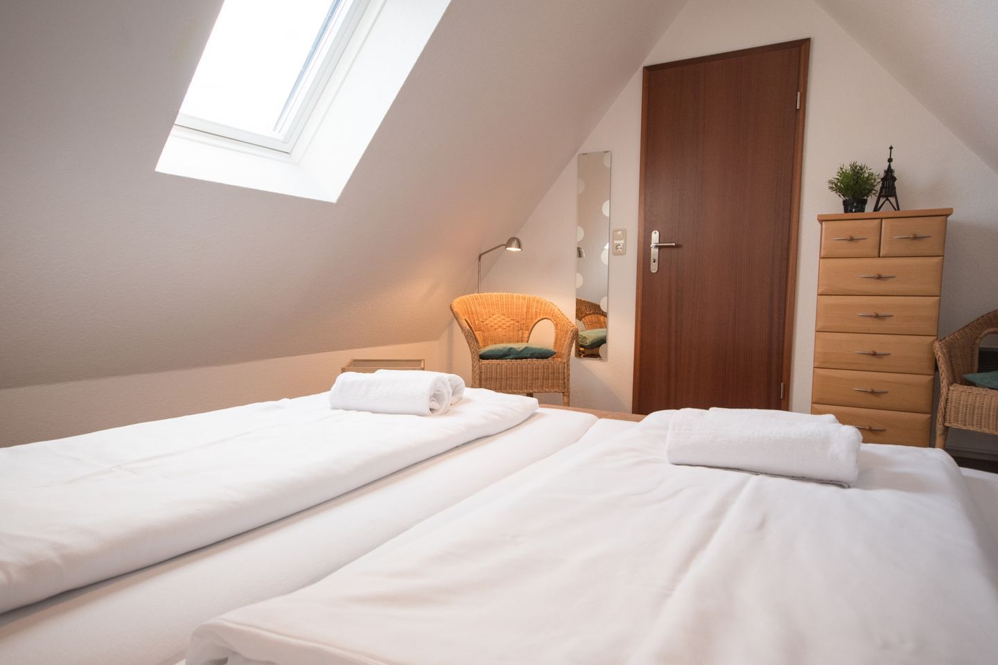 Ein Schlafzimmer mit zwei Betten, einem Holzschrank, einem Korbstuhl, einem kleinen Tisch und einem Dachfenster.