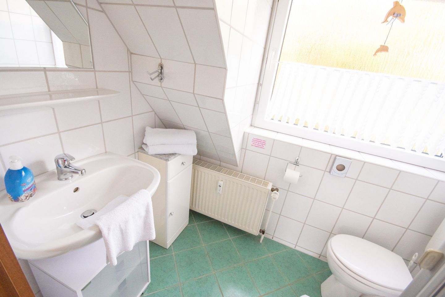 Ein Badezimmer mit einem weißen Waschbecken, einem Spiegel, einem Handtuchhalter, einer Toilette und einem Fenster mit Blick auf einen Zaun.