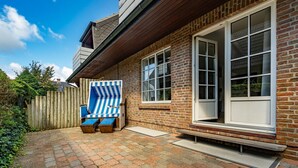 A brick house with a wooden deck, a large window, and a blue and white striped chair. - Wanlik Hüs I - 3-room vacation apartment, for max. 3 persons with terrace. (Sylt)