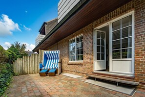 A brick house with a wooden deck, a large window, and a blue and white striped chair. - Wanlik Hüs I - 3-room vacation apartment, for max. 3 persons with terrace. (Sylt)