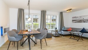 A modern living room with a wooden dining table, grey upholstered chairs, a grey sofa with colorful cushions, and a large window with curtains. - Wanlik Hüs I - 3-room vacation apartment, for max. 3 persons with terrace. (Sylt)
