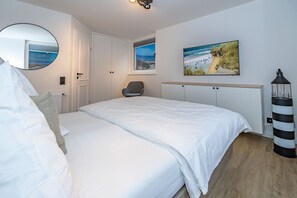 A bedroom with a bed, a chair, a desk, a lamp, and a painting of a beach scene. - Wanlik Hüs I - 3-room vacation apartment, for max. 3 persons with terrace. (Sylt)