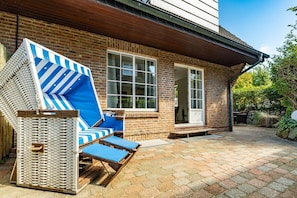 Une maison en briques avec un patio, deux fauteuils de jardin à rayures bleues et une table en bois.