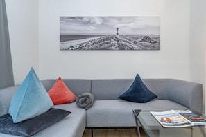 Un salon avec un canapé gris, trois coussins, une table basse en verre sur laquelle sont posés des magazines, et une photographie en noir et blanc d’un phare accrochée au mur.