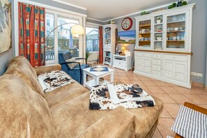 Uma sala de estar aconchegante com um sofá marrom, uma poltrona azul e uma mesa de centro branca. Há um armário branco com portas de vidro e um relógio na parede.