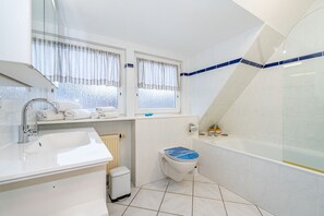 Ett modernt badrum med handfat, toalett och badkar. Det finns två fönster med gardiner, en handdukshängare och en liten papperskorg.
