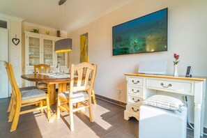 Un coin repas avec une table et des chaises en bois, une commode avec des tiroirs, un tableau accroché au mur et un bureau avec un ordinateur et un vase de fleurs.