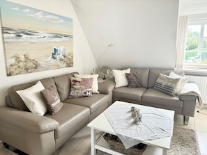 A modern living room with a beige leather sofa, a white coffee table, and a beach-themed painting on the wall. - Friesenglück - 3-room vacation apartment for up to 4 people with sunbathing area. (Sylt)
