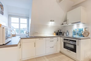 A modern kitchen with white cabinets, a built-in oven, and a large window providing natural light. - Friesenglück - 3-room vacation apartment for up to 4 people with sunbathing area. (Sylt)