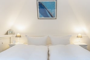 Een tweepersoonsbed met witte beddengoed, twee nachtkastjes met lampen, een ingelijste foto aan de muur en een klein horloge op een van de tafels.